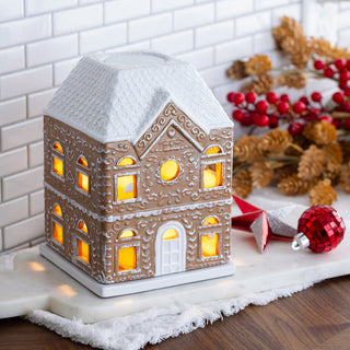 A gingerbread house-shaped ceramic lamp with a white snow-dusted roof and glowing warm yellow windows, styled on a white marble surface with holiday decorations in the background.