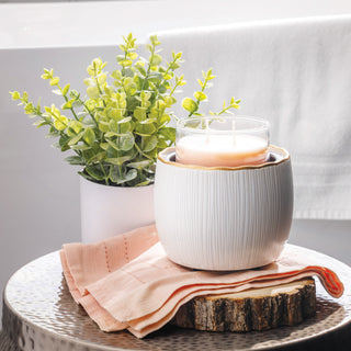 Candle in a textured white holder on a wooden coaster with a plant and pink napkin in the background.