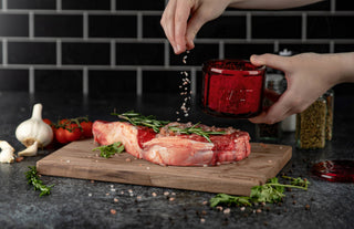 Person seasoning raw meat on a wooden cutting board with a black tiled wall in the background.